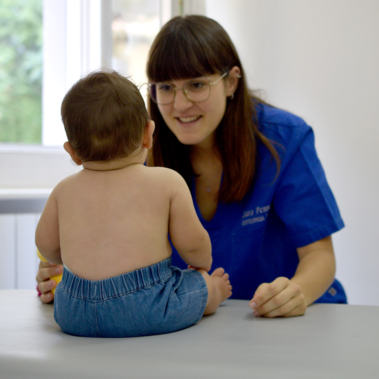 Osteopata Sara Penno durante una seduta di osteopatia pediatrica, sorridente con divisa blu con il suo nome ricamato nello studio luminoso, mentre osserva un neonato seduto sul lettino con pantaloncini in jeans. Ha capelli castani lisci con frangia a caschetto e porta occhiali con montatura sottile