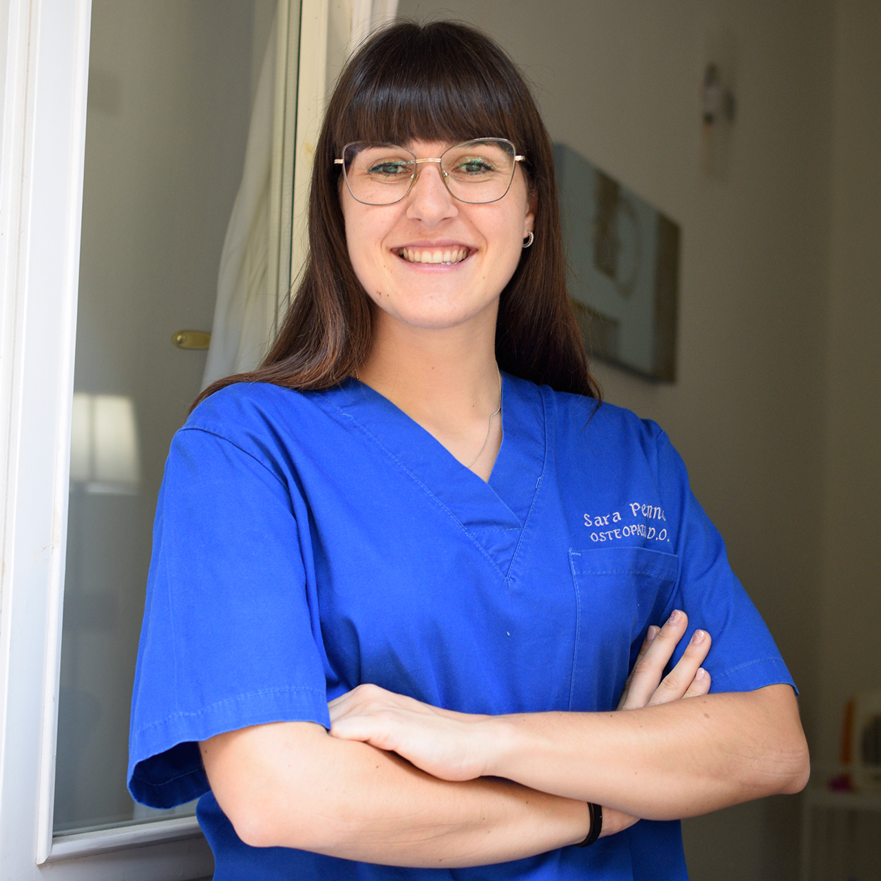 Osteopata Sara Penno posa sorridente all’ingresso del suo studio di osteopatia, con le braccia conserte. Indossa una casacca sanitaria blu con il suo nome ricamato, ha capelli castani lisci con frangia a caschetto e porta occhiali con montatura sottile. L’ambiente è luminoso e accogliente.