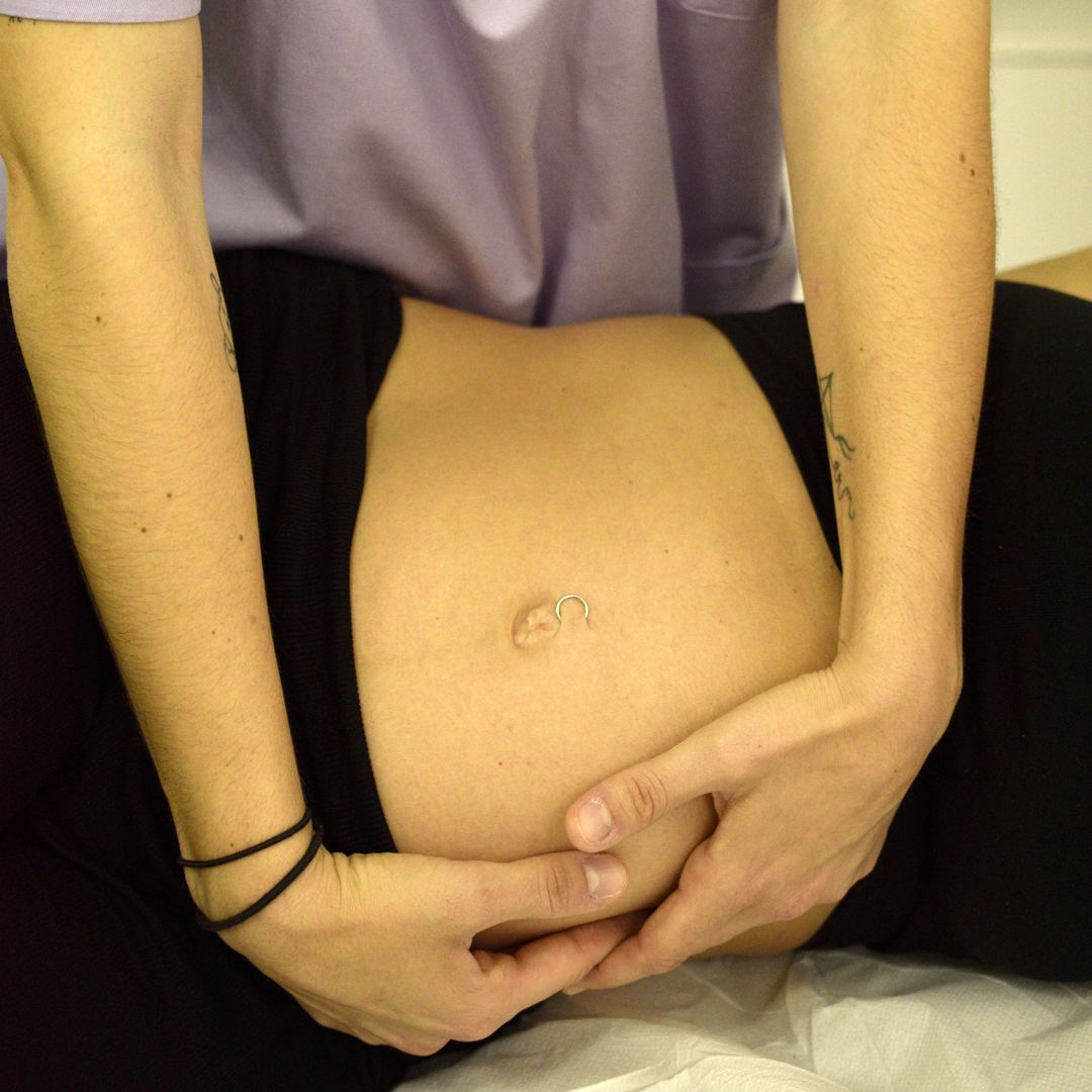 Osteopata Sara Penno durante una seduta di osteopatia in gravidanza esegue una manipolazione delicata sull’addome di una donna in gravidanza distesa su un lettino. Le mani dell’osteopata, con un tatuaggio a forma di aquilone sul braccio, sono posate sul ventre della paziente che indossa una maglietta nera. Indossa una casacca sanitaria color lilla e due elestici neri al polso destro. L’ambiente è quello dello studio medico.