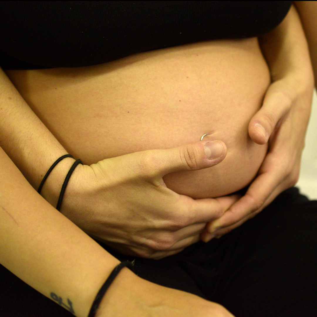 Osteopata Sara Penno durante una seduta di osteopatia in gravidanza esegue una manipolazione delicata sull’addome di una donna in gravidanza distesa su un lettino. Le mani dell’osteopata sono posate sul ventre della paziente che indossa una maglietta nera.
Al polso ha due elastici neri, mentre la paziente un elsatico nero al polso, un piercing all'ombelico e un tatuaggio di un ancora. L’ambiente è quello dello studio medico.