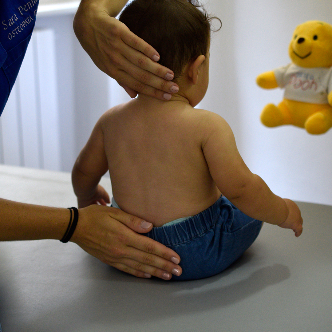 Osteopata Sara Penno durante una seduta di osteopatia pediatrica esegue una delicata valutazione sul bambino, appoggiando una mano sulla nuca e l’altra sulla zona lombare. Il bambino è seduto a dorso nudo su un lettino grigio, indossa pantaloni in denim chiaro, mentre sullo sfondo si intravede un peluche giallo di Winnie the Pooh. L’osteopata indossa una casacca sanitaria blu con il suo nome ricamato.