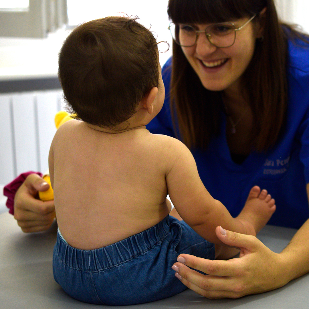 Osteopata Sara Penno durante una seduta di osteopatia pediatrica, sorridente con divisa blu con il suo nome ricamato nello studio luminoso, mentre osserva un neonato seduto sul lettino con pantaloncini in jeans. Ha capelli castani lisci con frangia a caschetto e porta occhiali con montatura sottile