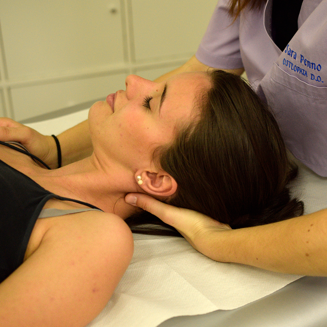Osteopata Sara Penno durante una seduta di osteopatia esegue una manipolazione delicata al collo di una paziente distesa su un lettino bianco, nello studio medico. L’osteopata indossa una casacca sanitaria color lilla con il suo nome ricamato e al polso destro un elastico nero, mentre la paziente porta una canotta nera, la capelli castani legati da una coda e un orecchino bianco. l'osteopatia