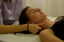 Osteopata Sara Penno durante una seduta di osteopatia esegue una manipolazione delicata al collo (dolore Cervicale) di una paziente distesa su un lettino bianco, nello studio medico. L’osteopata indossa una casacca sanitaria color lilla con il suo nome ricamato e al polso destro un elastico nero, mentre la paziente porta una canotta nera, la capelli castani legati da una coda e un orecchino bianco. l'osteopatia