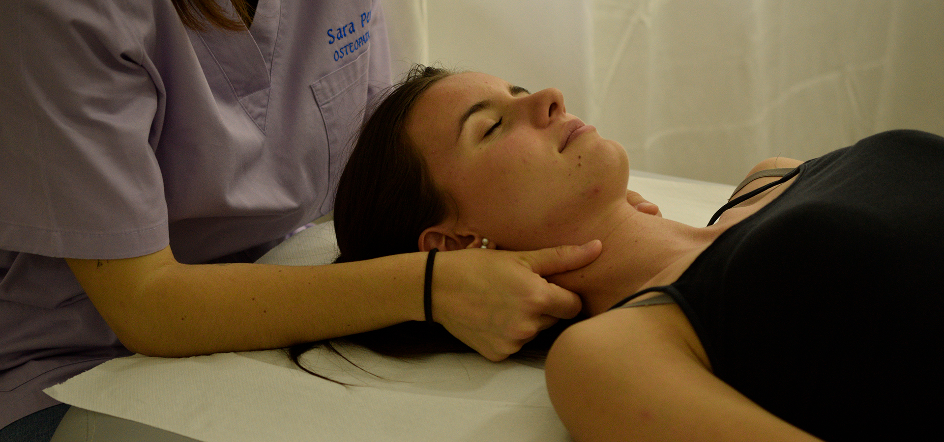 Osteopata Sara Penno durante una seduta di osteopatia esegue una manipolazione delicata al collo di una paziente distesa su un lettino bianco, nello studio medico. L’osteopata indossa una casacca sanitaria color lilla con il suo nome ricamato e al polso destro un elastico nero, mentre la paziente porta una canotta nera, la capelli castani legati da una coda e un orecchino bianco