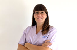 Osteopata Sara Penno posa sorridente nel suo studio di osteopatia, con le braccia conserte. Indossa una casacca sanitaria lilla con il suo nome ricamato, ha capelli castani lisci con frangia a caschetto e porta occhiali con montatura sottile. L’ambiente è luminoso e accogliente. Che cos'è l'osteopatia? Perché è affidarsi a un osteopata?