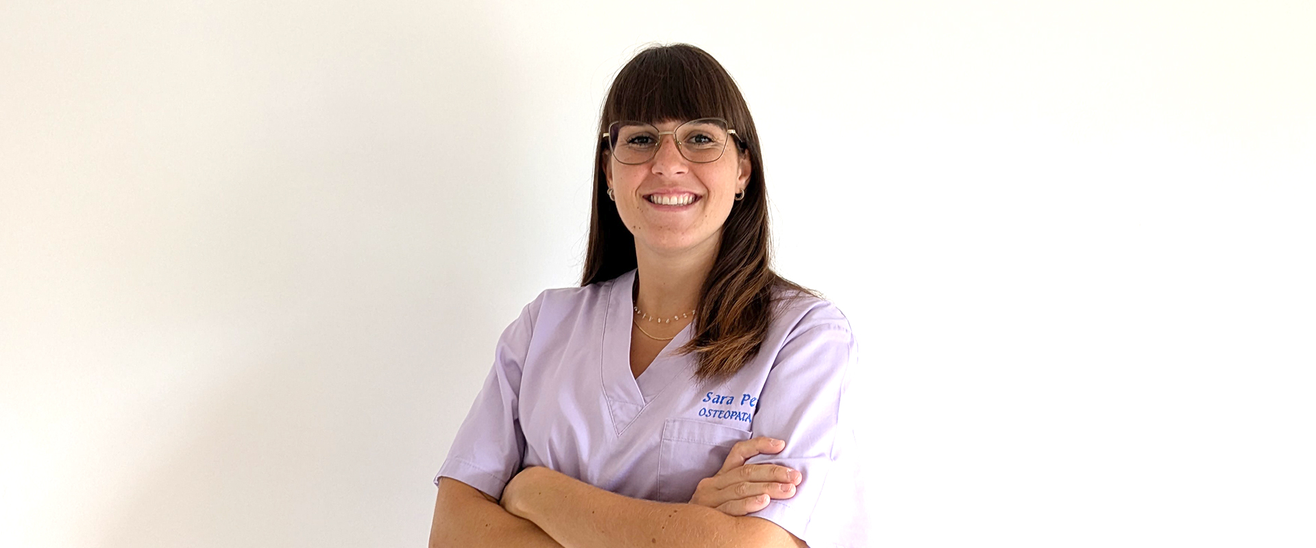 Osteopata Sara Penno posa sorridente nel suo studio di osteopatia, con le braccia conserte. Indossa una casacca sanitaria lilla con il suo nome ricamato, ha capelli castani lisci con frangia a caschetto e porta occhiali con montatura sottile. L’ambiente è luminoso e accogliente. Che cos'è l'osteopatia? Perché è affidarsi a un osteopata?