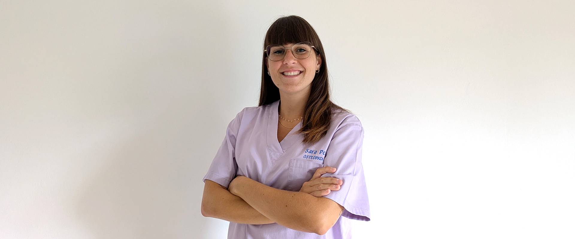Osteopata Sara Penno posa sorridente all’ingresso del suo studio di osteopatia. Indossa una casacca sanitaria blu con il suo nome ricamato, ha capelli castani lisci con frangia a caschetto e porta occhiali con montatura sottile. L’ambiente è luminoso e accogliente.